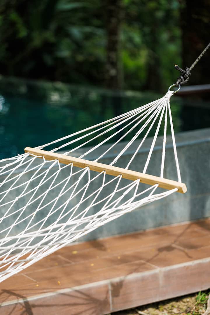 A close-up of a white hammock with wooden supports, positioned near a pool surrounded by lush greenery at Templer Park in Rawang.