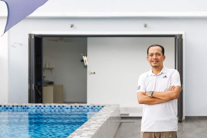 A modern backyard view featuring a tiled swimming pool and a white open-door building in Selangor, with a person standing confidently beside the pool.