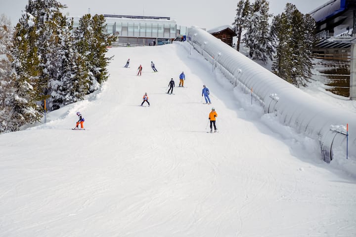 Des skieurs descendent une pente enneigée entourée de pins et de bâtiments modernes, illustrant les opportunités en matière de tourisme et d'activités de loisirs pour les villes hôtes.