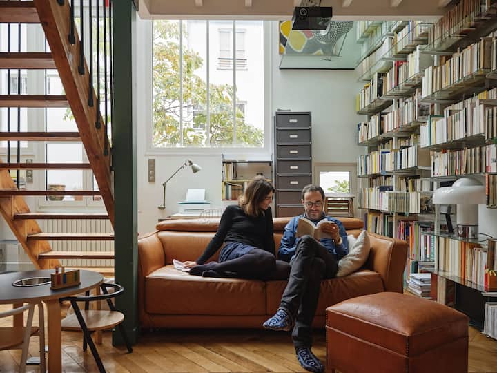L'intérieur lumineux et accueillant d'une pièce de vie avec un canapé en cuir marron, de grandes étagères murales remplies de livres et de grandes fenêtres donnant sur un arbre à l'extérieur, reflétant les options d'hébergement durables.