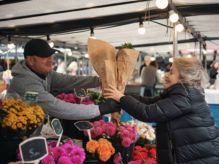 Un fleuriste remet un bouquet emballé à une cliente sur un étal de marché en plein air animé, rempli de fleurs colorées et d'étiquettes de prix.