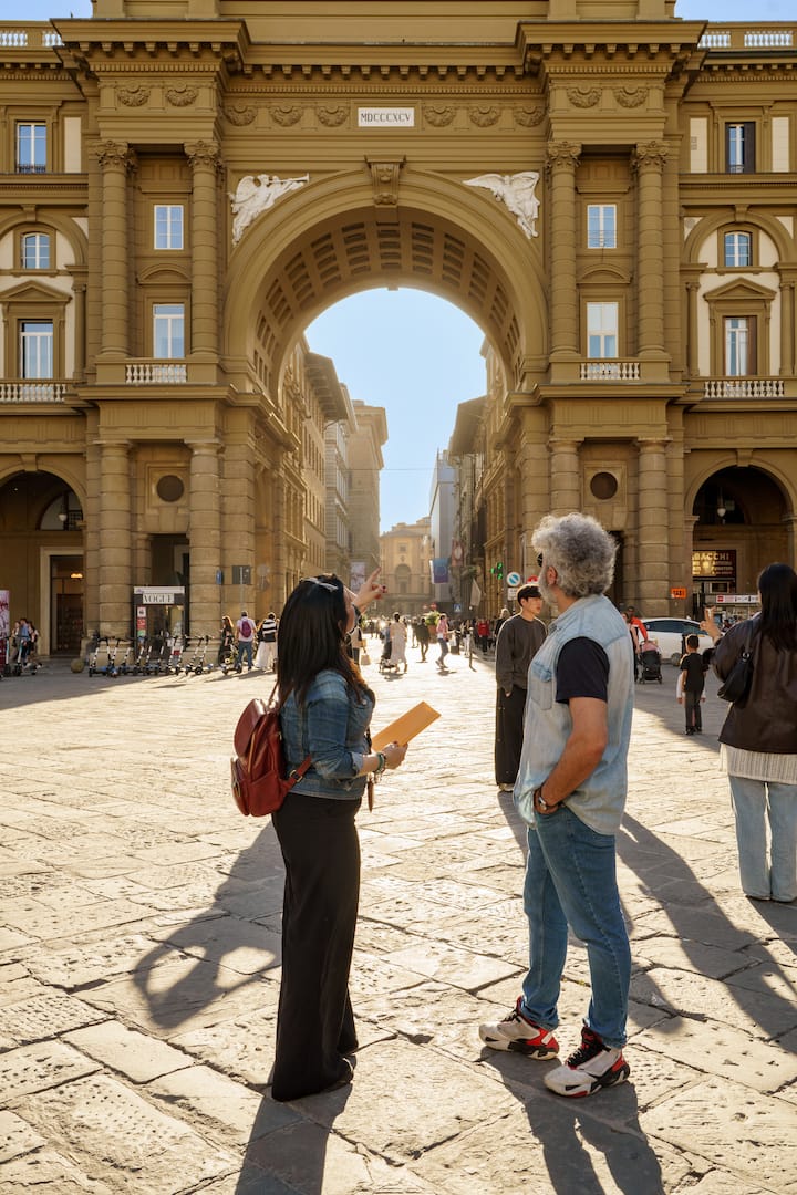 Un centre historique peuplé de piétons avec une grande arche devant laquelle deux personnes s'entretiennent.