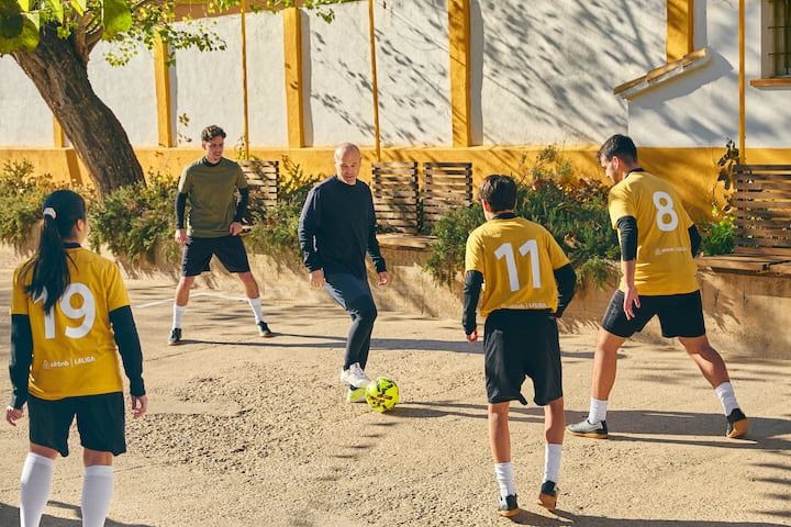 Un grupo de personas juega al fútbol en un patio soleado. Algunos de los jugadores llevan camisetas amarillas con los números 19, 11 y 8. El balón está colocado cerca del centro.