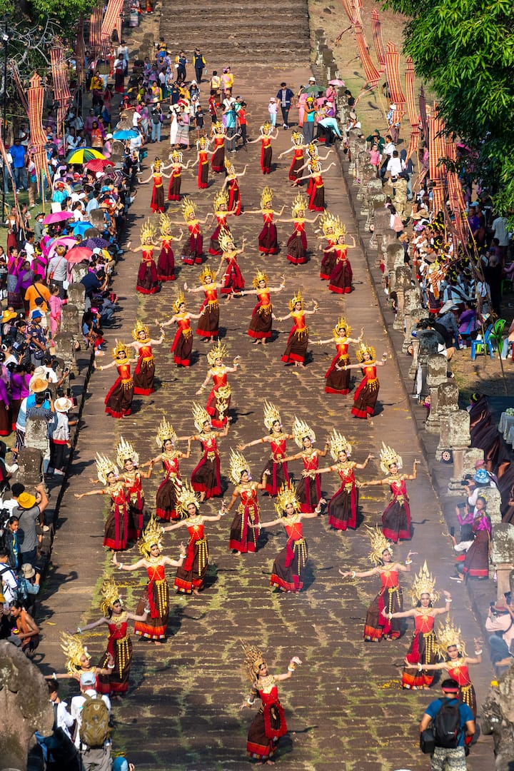 A vibrant cultural festival in buriram featuring a group of dancers in ornate gold and red traditional attire performing on a historic stone pathway, surrounded by spectators under colorful umbrellas.