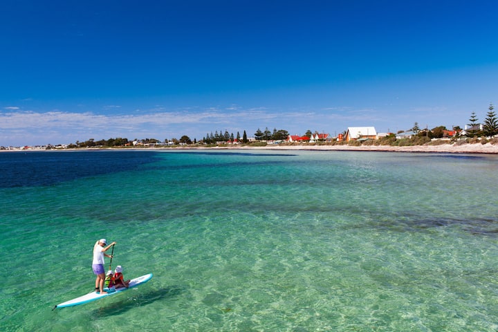 A serene coastal scene with turquoise water, a person paddleboarding with a child, and a distant shoreline featuring houses and trees under a clear blue sky.
