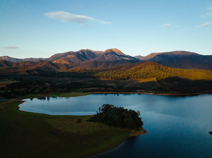 A tranquil rural landscape at sunset featuring a calm lake surrounded by green fields and forested mountains in the background.