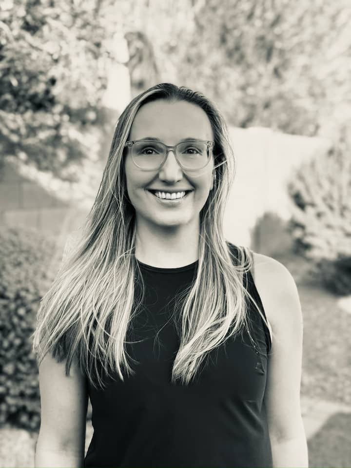 Black and white photo of Selma standing in a black tank top, wearing glasses in front of a garden landscape 