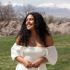 Image of a woman wearing a white top, long black curly hair, standing in front of a mountain range smiling in the sun