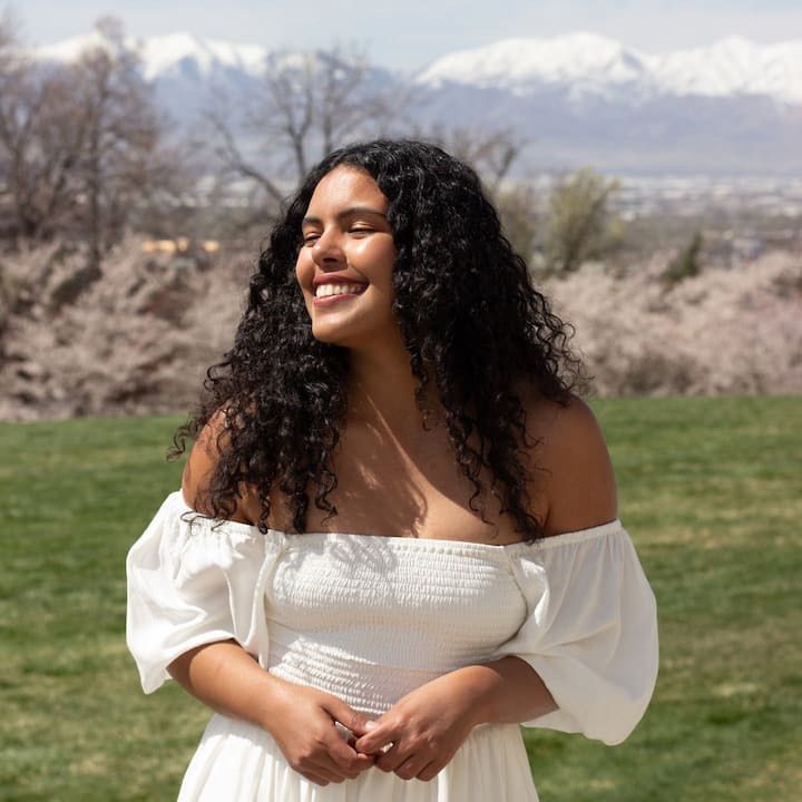Image of a woman wearing a white top, long black curly hair, standing in front of a mountain range smiling in the sun