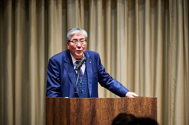 A person stands at a wooden podium, speaking into a microphone, with beige curtains in the background, during a formal event focused on community and tourism collaboration in Osaka.