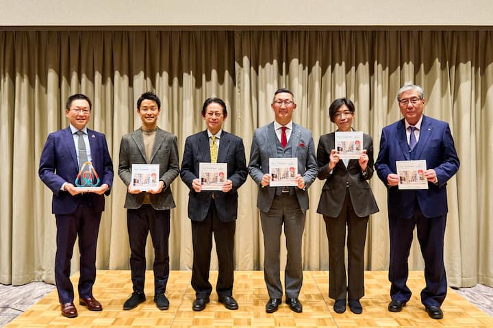 A group of six individuals standing in a formal setting, holding documents related to Airbnb Japan's public policy initiatives. A curtain backdrop is visible behind them.
