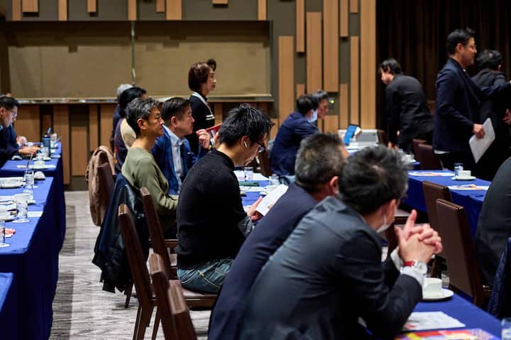 Attendees seated at a formal meeting in Japan, with blue tablecloths and materials on the tables in a modern conference room setting.