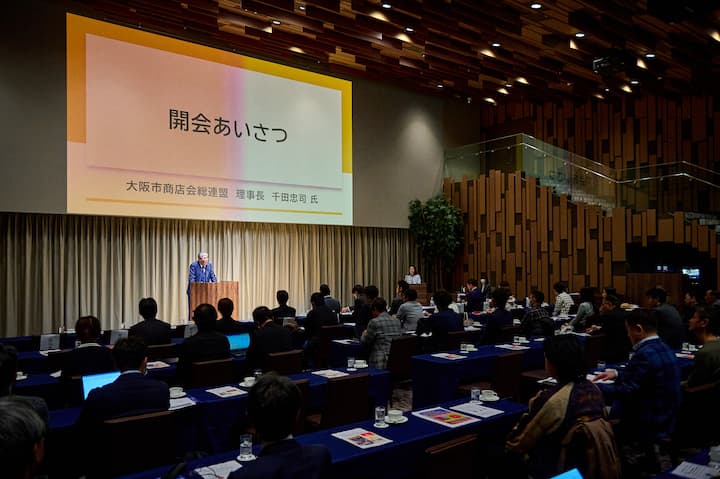 A speaker at a podium addressing an audience in a conference room. A large presentation screen above displays Japanese text, including '開会あいさつ' (opening greeting).