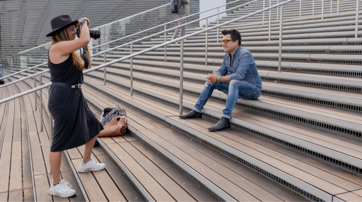 Une photographe portant une robe noire et un chapeau prend en photo une personne assise sur des escaliers modernes en bois, mettant l'accent sur l'interaction et les instructions.