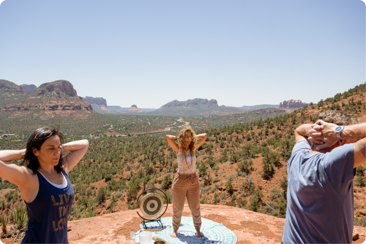 Des participants s'étirent sur un plateau rocheux. Un gong et des rafraîchissements sont disposés sur un tapis, surplombant une vaste vallée désertique parsemée de formations rocheuses à l'horizon.