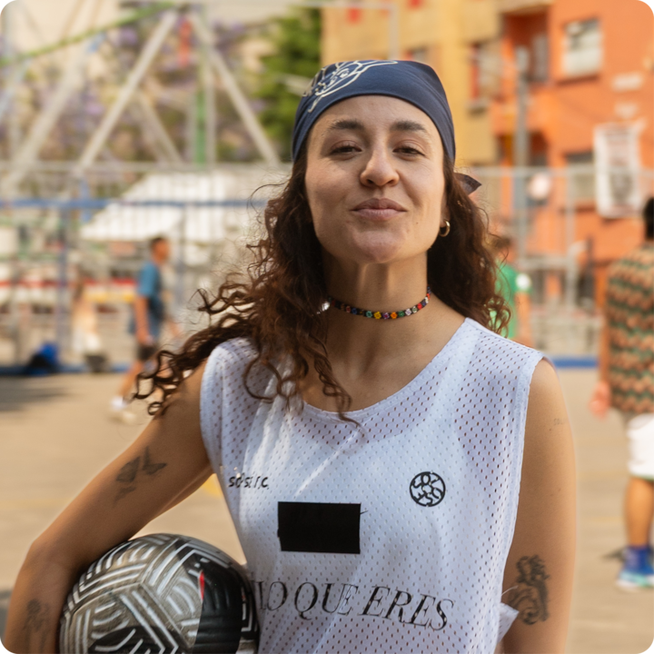 Une personne vêtue d'une tenue de sport tient un ballon de football aux motifs complexes. Elle porte un haut en maille blanche avec du texte, un collier coloré et un bandana. En arrière-plan, on aperçoit des bâtiments et une zone clôturée.
