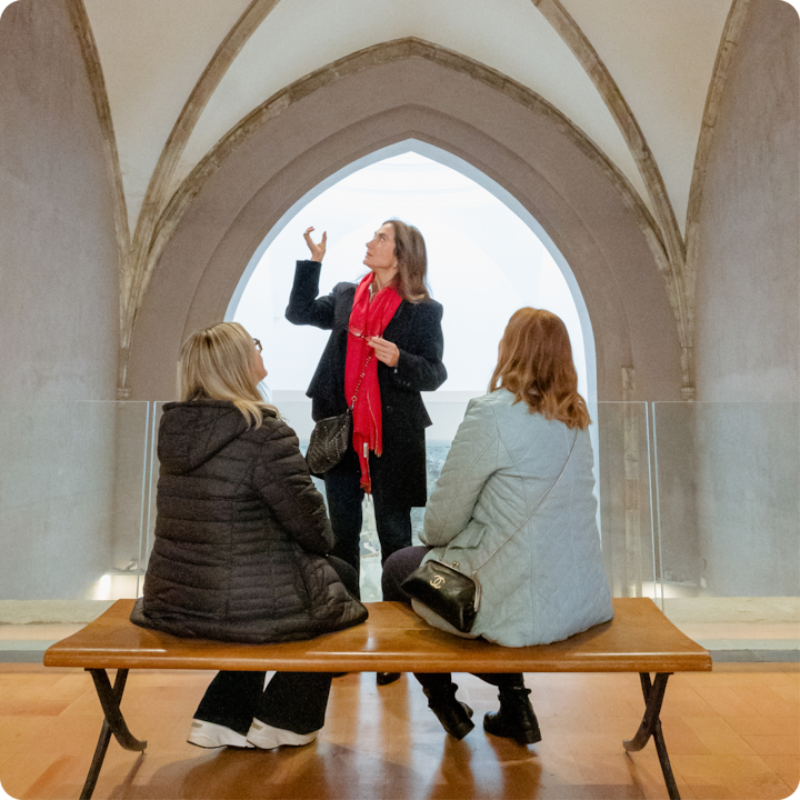 Une personne fait des gestes tout en discutant avec deux participants dans un espace historique encadré par une grande arche en pierre baignée de lumière naturelle.