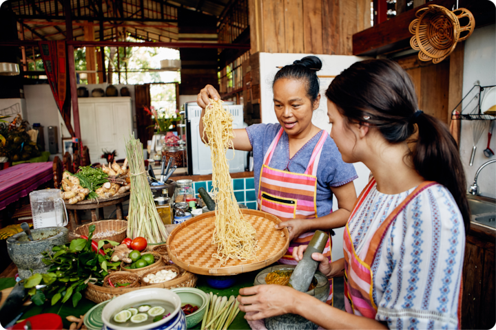 Deux personnes préparent un plat de nouilles traditionnel dans une cuisine rustique, entourées d'ingrédients frais comme du citron vert, du piment et des herbes sur un comptoir en bois.