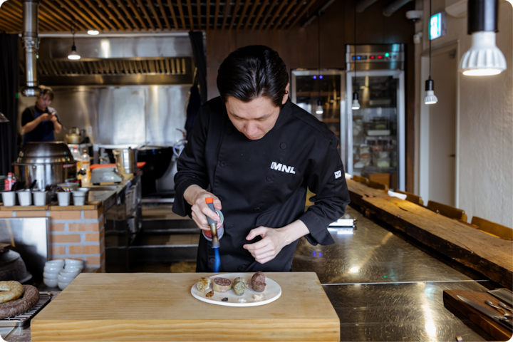 Une cuisine professionnelle où un chef prépare un plat à l'aide d'un chalumeau de cuisine. Le plan de travail est garni de divers ustensiles de cuisine.