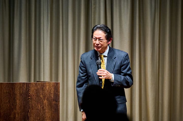 A professional presenter in a suit giving a speech on stage, holding a microphone near a podium, with beige curtains forming the background, related to the topic of '住んでよし、訪れてよし、稼いでよし'.