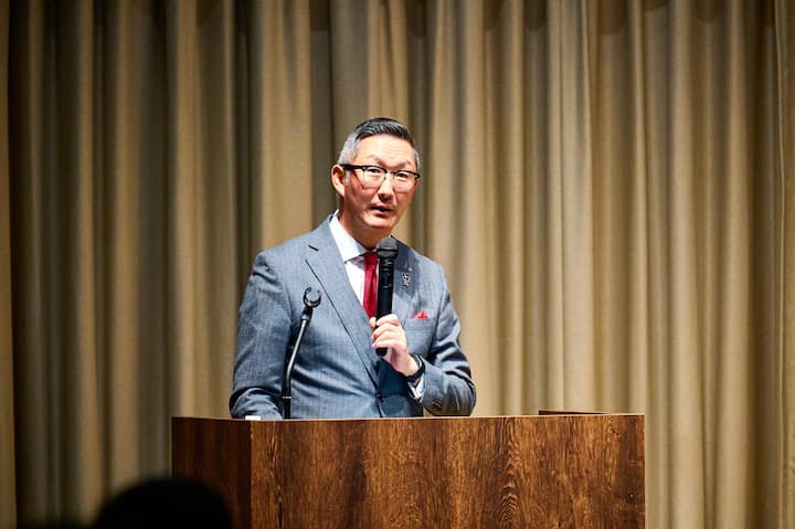 A person in formal attire stands at a wooden podium, holding a microphone, with beige curtains in the background, delivering a presentation.