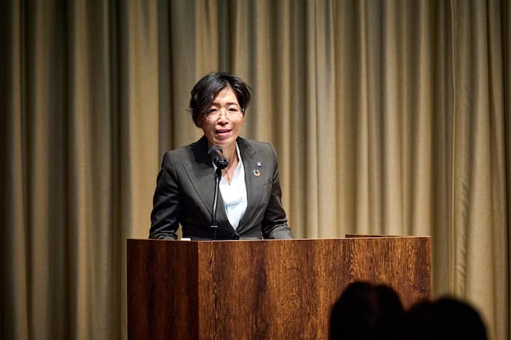 A person gives a presentation at a podium with beige curtains behind them, wearing a dark blazer with a sustainable development pin.