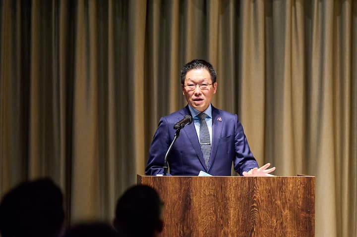 A person in a navy suit speaks at a podium during an event, with a microphone positioned on the podium and beige curtains in the background.