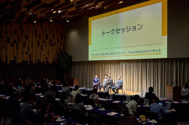A panel discussion taking place in a modern conference room with four speakers seated on stage under a large screen displaying Japanese text that reads 'トークセッション' and additional text about community revitalization efforts.