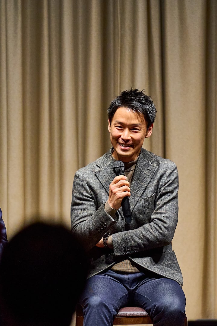 A person wearing a gray blazer and holding a microphone, seated on a chair in front of beige curtains during a panel discussion.