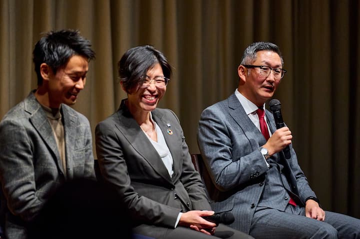 Three individuals seated on a stage, dressed in formal attire, with one holding a microphone, participating in a panel discussion against a beige curtain backdrop.