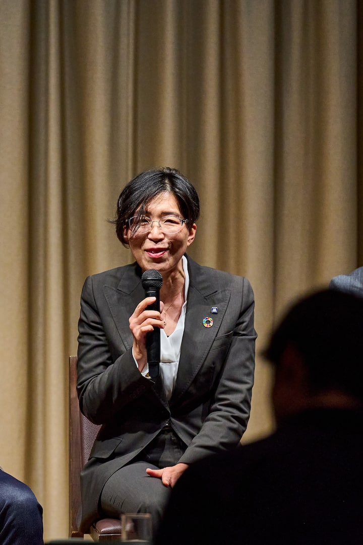 An individual speaking into a microphone while seated, wearing a dark suit and a pin representing the Sustainable Development Goals, with beige curtains in the background.