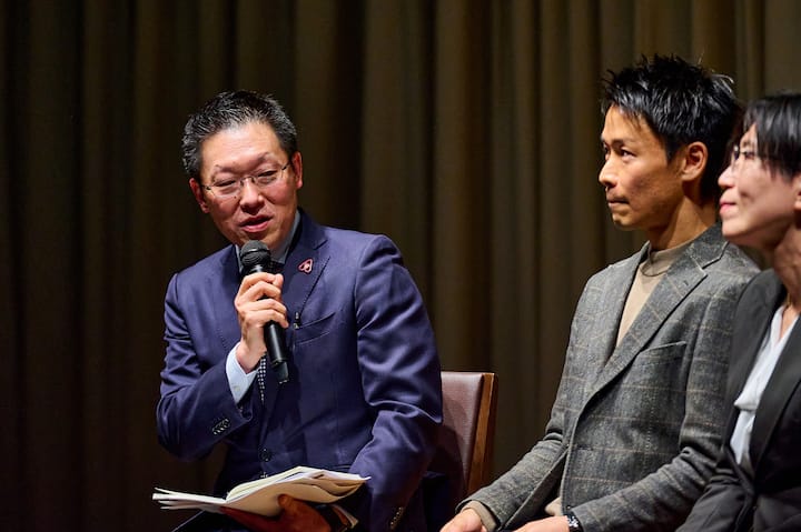 A panel discussion featuring individuals seated, one holding a microphone and notebook, against a curtain backdrop.
