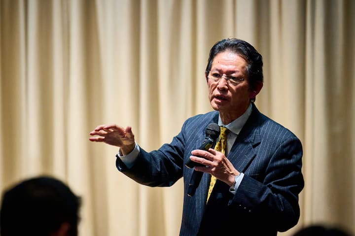 A person in a suit holding a microphone and gesturing with one hand against a backdrop of beige curtains, suggesting a formal presentation or speech.