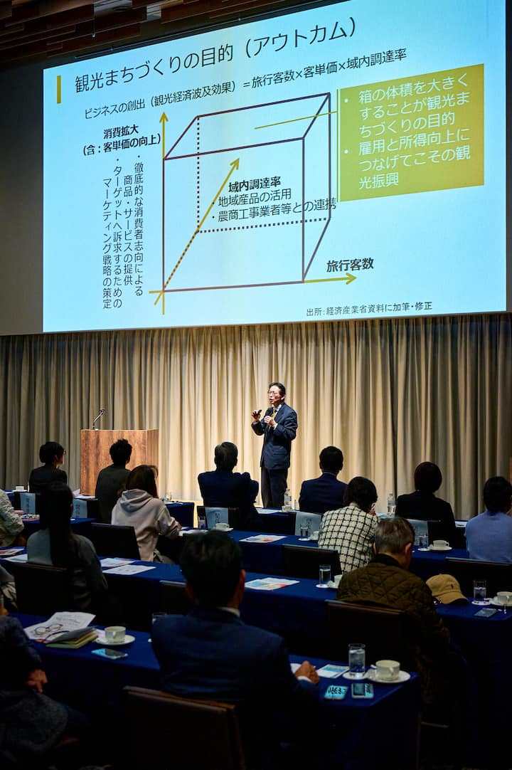 A man in a suit stands on stage giving a presentation about tourism policy in Japan, with a large screen displaying a diagram and Japanese text about creating tourism strategies.