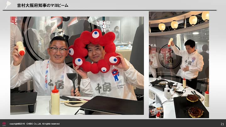 Two people at a cooking station, one holding a red, eye-themed plush wreath and the other holding a bottle of mayonnaise, with griddles and cooking tools visible in front of them.
