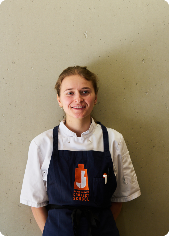 Gros plan sur la veste blanche et le tablier bleu foncé d'une cheffe, avec un stylo dans la poche du tablier.