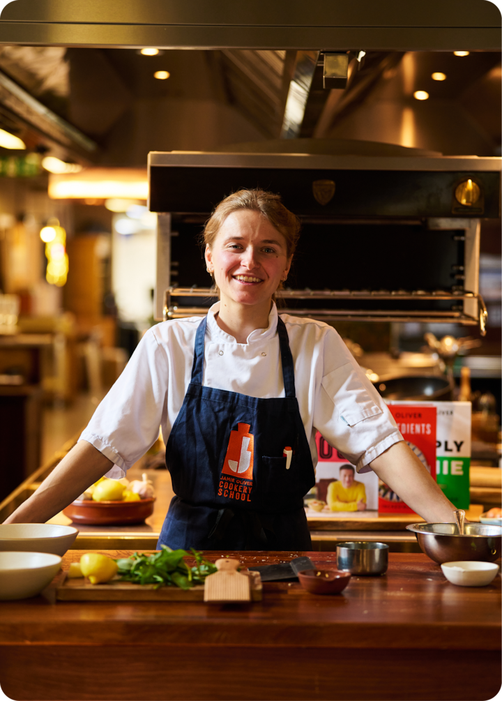 Une cuisine avec une cheffe en tablier bleu marine. Le plan de travail présente des produits frais, des petits bols et des ustensiles de cuisine, suggérant un cours de cuisine ou une préparation culinaire.