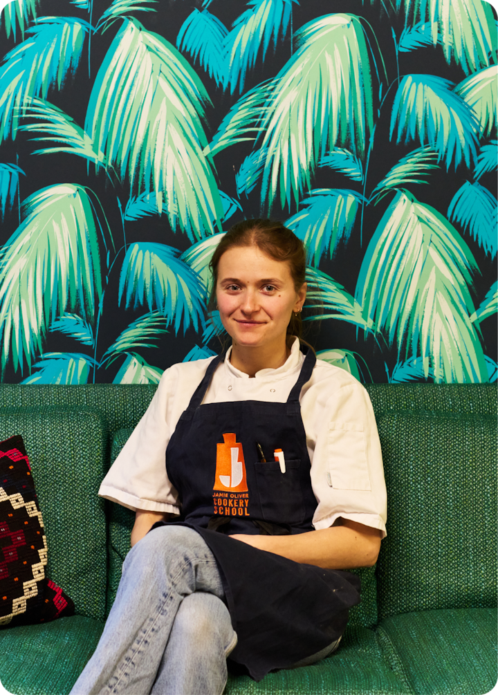 Une personne portant un tablier bleu foncé est assise sur un canapé vert, devant un papier peint à feuilles de palmier éclatantes.