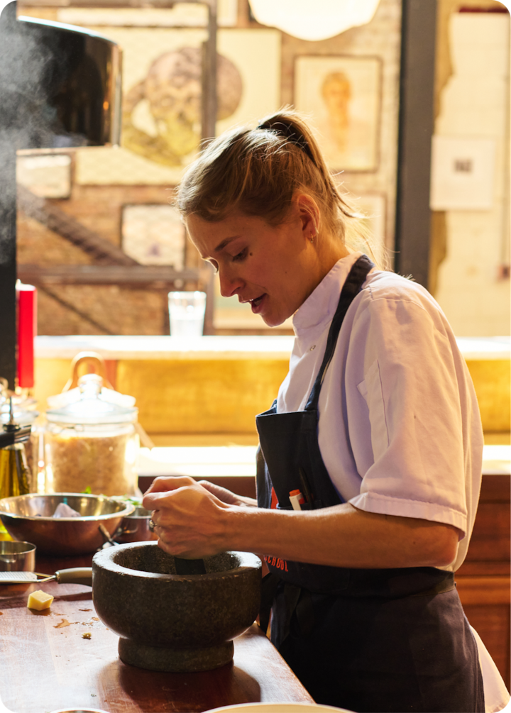 Une scène culinaire montrant une personne qui écrase des ingrédients dans un mortier en pierre avec un pilon, dans une ambiance de cuisine conviviale et avec des œuvres d'art encadrées en arrière-plan.