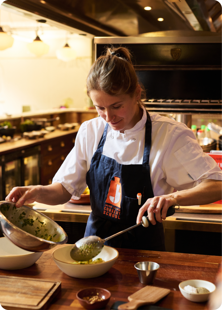 Gros plan sur une cheffe qui prépare un plat avec des herbes fraîches dans une cuisine professionnelle. Les ustensiles et les ingrédients sont disposés sur un plan de travail en bois.