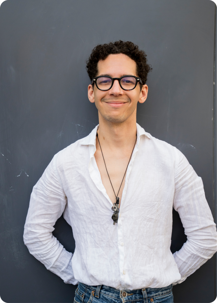 Une personne se tient debout, les mains sur les hanches, sur un fond bleu foncé. Elle porte une chemise blanche et un jean bleu.