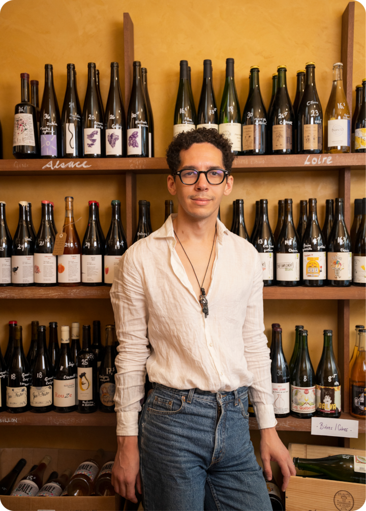 Une personne se tient debout devant des étagères en bois remplies de bouteilles de vin, étiquetées de notes manuscrites, dans une pièce aux tons chauds.