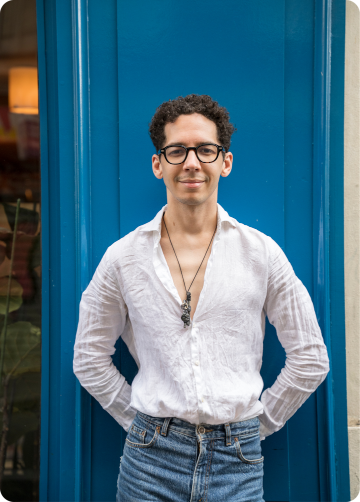 Un homme s'appuie contre une porte bleu vif. Il porte une chemise blanche décontractée, un jean bleu et un collier avec un pendentif original.