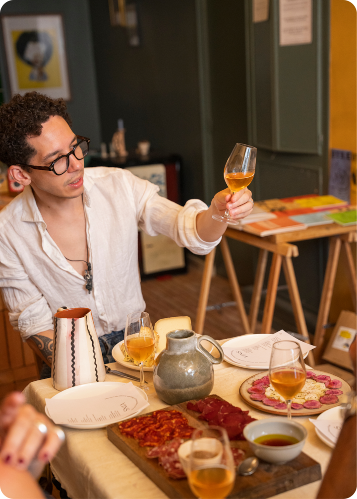 Une table dressée avec de la charcuterie, des fromages et des verres de vin orange dans une salle à manger conviviale au décor rustique. Une personne assise à table lève un verre de vin pour porter un toast.