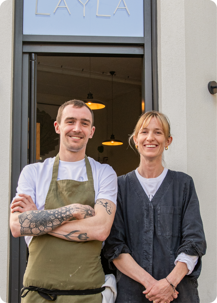 Deux hôtes se tiennent devant un établissement dont le nom « Layla » est affiché à l'extérieur, au-dessus de la porte.