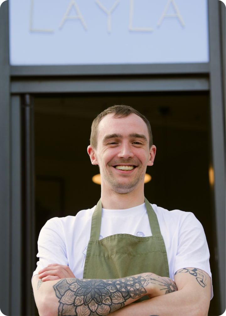 Une personne portant un tablier vert et une chemise blanche se tient avec assurance devant un espace portant l'inscription « Layla ». Ses bras tatoués sont croisés.