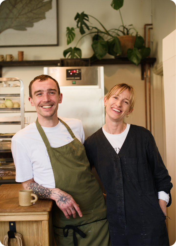 Une cuisine accueillante avec deux hôtes, dont l'un est appuyé contre un comptoir en bois sur lequel repose une tasse. Les hôtes sont entourés de plantes, d'un réfrigérateur et de pâtisseries.