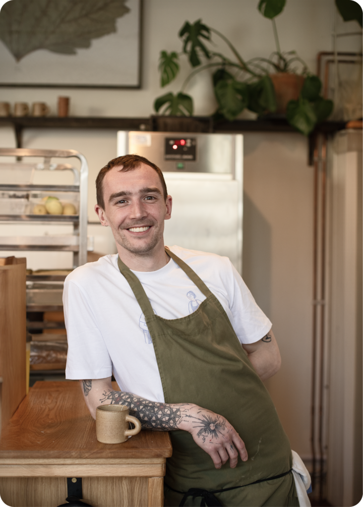 Une personne en tablier vert et tenue décontractée s'appuie sur un comptoir en bois dans une cuisine avec des plantes retombantes, des étagères et un réfrigérateur en arrière-plan.