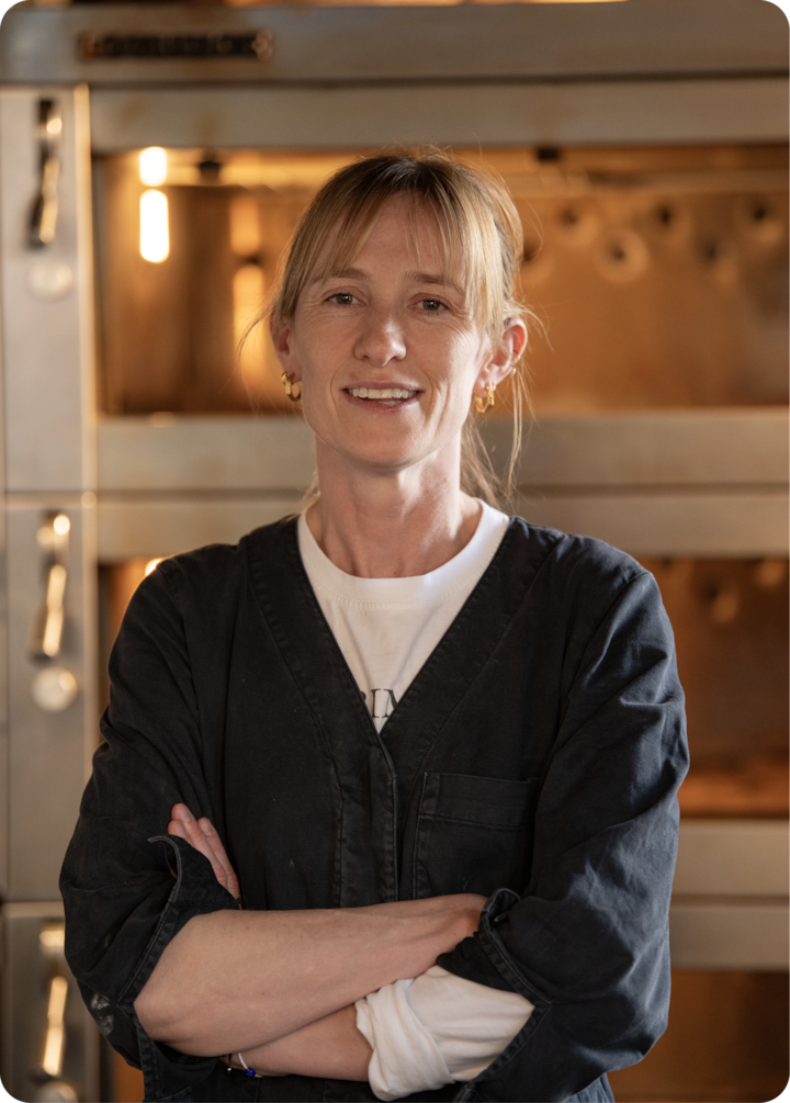 Une personne, les bras croisés devant un four professionnel, portant une veste sombre et une chemise blanche, se tient dans une cuisine accueillante et bien éclairée.