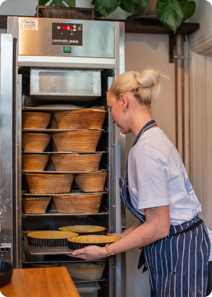 Une boulangère en tablier rayé glisse une plaque de fonds de tarte crus dans un four, entourée de bannetons ; des plantes et des décorations de cuisine sont visibles en arrière-plan.